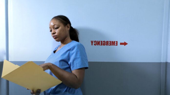 A nurse in blue scrubs reading a patient chart in a hospital corridor.